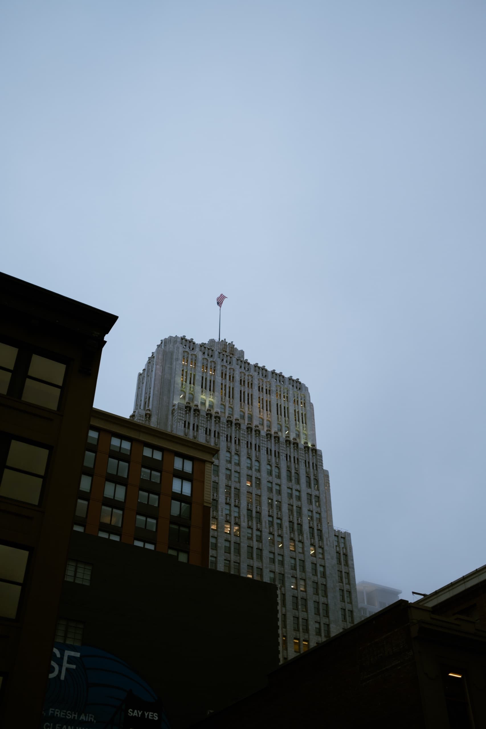 Looking up at an Art Deco stone tower and flag under a foggy sky.