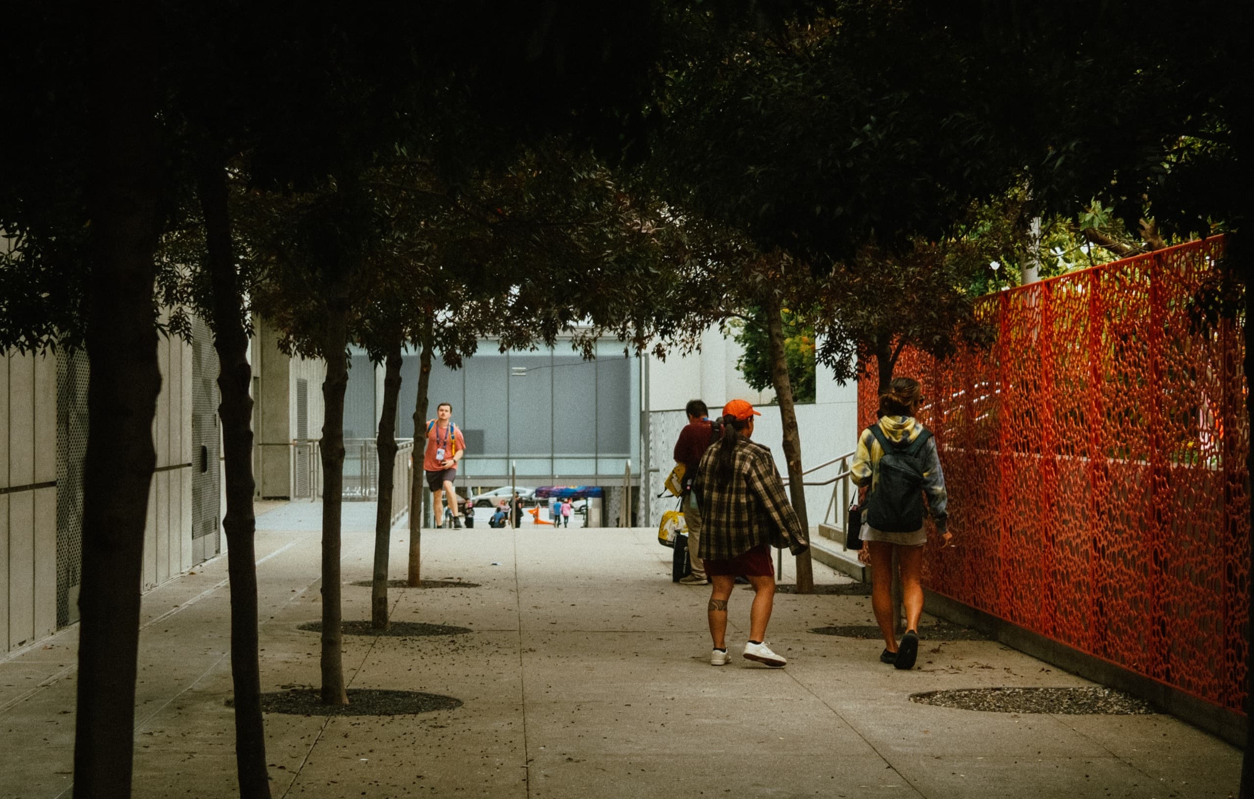 Walkers on a tree-lined Salesforce Park path beside a tall orange latticed fence.